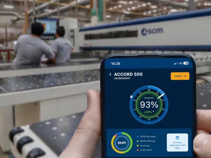 The SCM Digital Portal companion on a phone held on the factory floor, showing a machine at 93% productivity, with machines and a worker blurred behind it