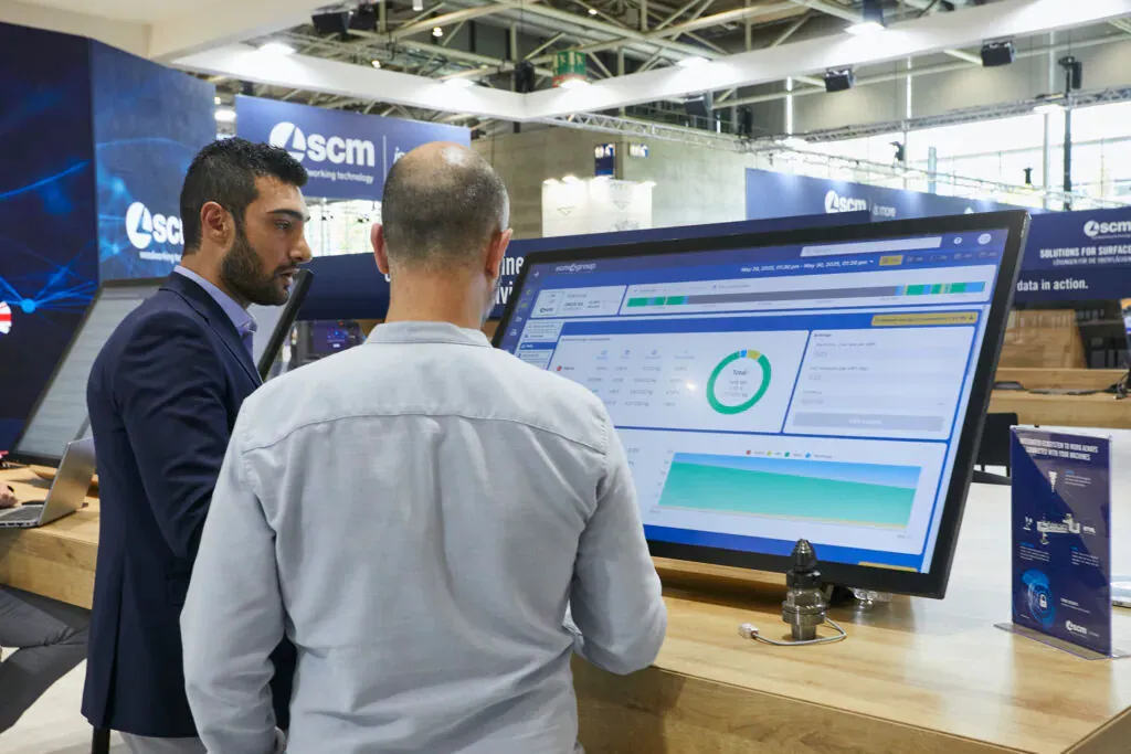 SCM Digital Portal running on a large monitor at SCM’s digital-services stand at a trade fair, a visitor and an SCM engineer in front of it